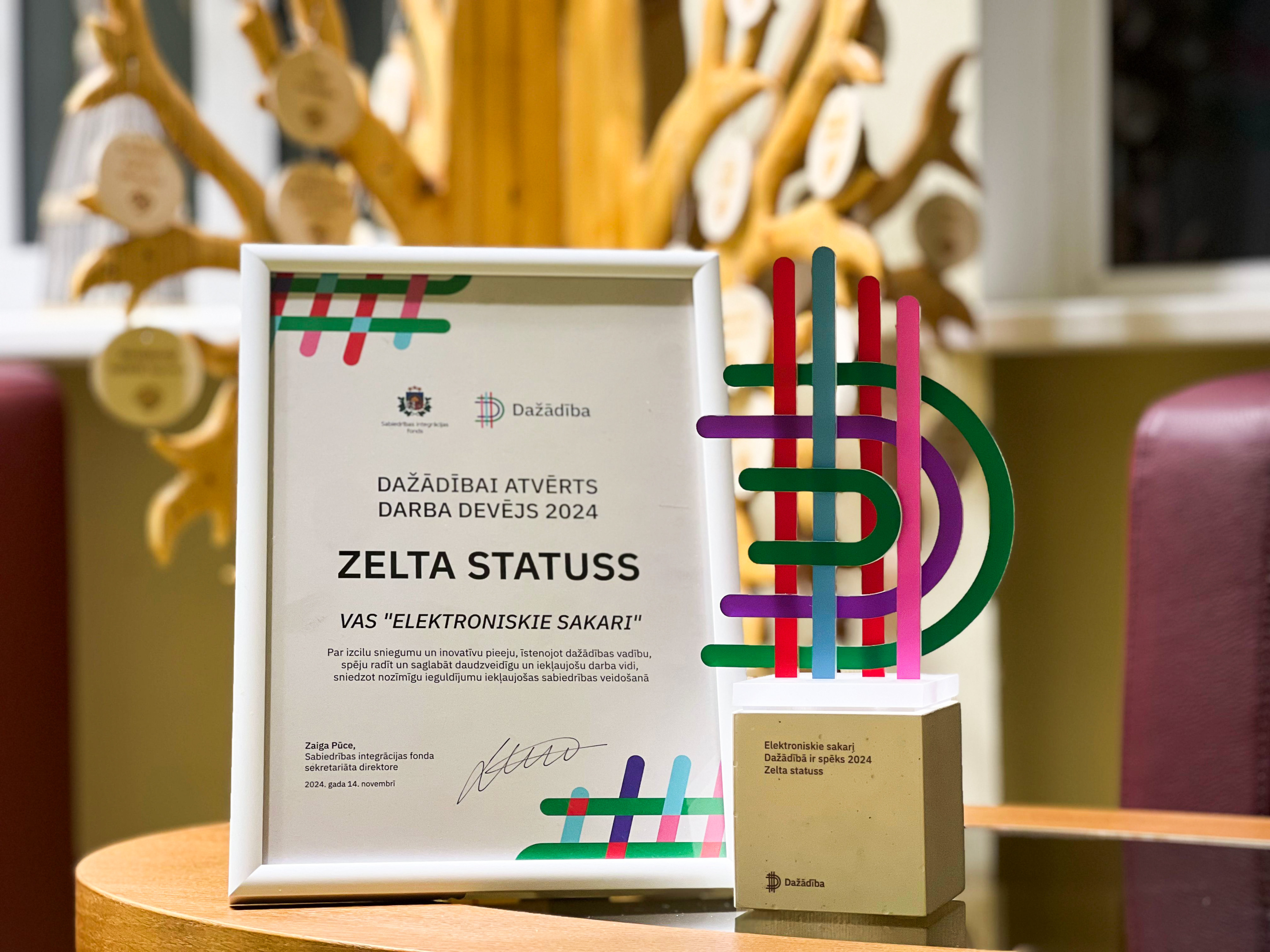 Fotogrāfija ar apliecinājumu un trofeju, kas piešķirti VAS 'Elektroniskie sakari' par Zelta statusu programmā 'Dažādībai atvērts darba devējs 2024'. Rāmis satur tekstu par uzņēmuma sasniegumiem daudzveidības un iekļaujošas sabiedrības veidošanā, un fonā redzams koka dekors ar simboliskiem elementiem.